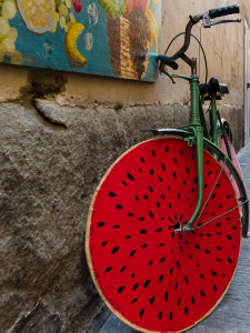 Against a city wall leans a green bicycle whose front wheel looks like a cross section of watermolon